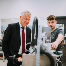 Person in a business suit and another in work attire examining a technical system with cables and control components.