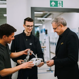 Two young individuals presenting a model car with an attached robotic arm to another person in a modern training environment.