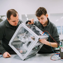 Two individuals working on an open metal frame containing mounted components and wiring, focused activity in a workshop setting.